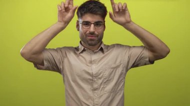 Gözlüklü İspanyol genç adam parlak yeşil stüdyoda taş boynuzları hareketi yapıyor; oyunculuk enerjisi.