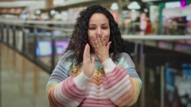 Renkli kazaklı genç İspanyol kadın alışveriş merkezinin hareketli merkezinde dikilirken ağzını neşeli bir ifadeyle kapatıyor. Etrafı dükkanlar ve canlı ışıklarla çevrili..