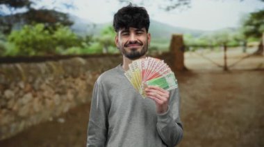 Parkta Norveçli banknotları tutan yakışıklı bir adam. Etrafı doğayla çevrili güvenle gülümsüyor, finans ve yaşam tarzı unsurlarını yaratıcı bir şekilde harmanlıyor..