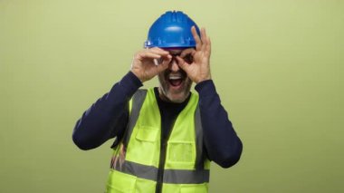 Mavi kask takan ve sarı yelek giyen bir adam yeşil arka planda şakacı hareketler sergiliyor.