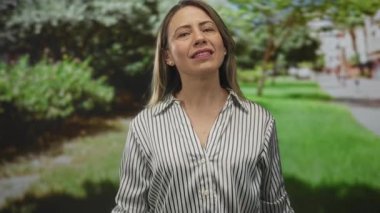 Çizgili bluzlu kadın yeşil çimlerin yanındaki park ağaçlarının altında el işareti yapıyor; dinginlik farkındalığı dengesi.