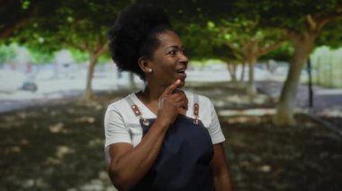 Önlük giymiş, gülümseyen ve yemyeşil bir parkta duran ağaçların arasından süzülen benekli güneş ışığıyla sıcak bir açık hava yaratan bir kadın..