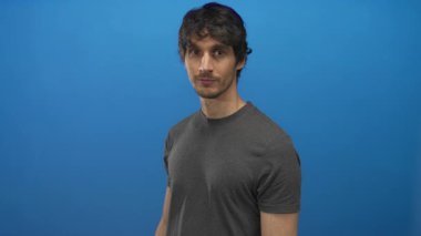 Genç adam parlak mavi bir stüdyo ortamında kolları çapraz gri bir gömlek içinde gülümsüyor..
