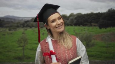 Mezuniyet şapkalı genç bir kadın elinde diploma ve kitap, manzaralı bir park ortamında gülümsüyor, açık havada akademik başarıyı kutluyor..