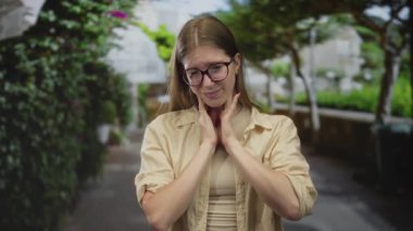 Güneş ışığını yansıtan gözlüklü, bej gömlekli, taş bina yakınında boyuna dokunan genç sarışın kadın. Stres kaygısı..