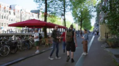 Güneşli bir Avrupa caddesinde ağaçlar, bisikletler ve arka planda kırmızı kafe şemsiyeleriyle yürüyen insanlar..