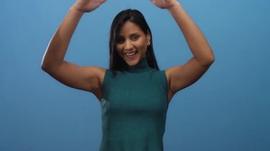 İzole edilmiş mavi bir arka plana karşı ellerini başının üstüne koyan genç Latin kadın neşeli ve modern bir ortama duyduğu güveni ifade ediyor..