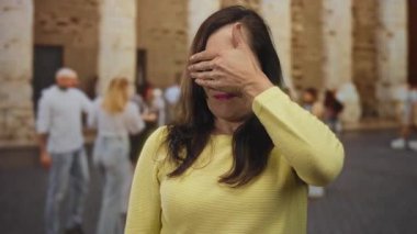 Tarihi caddede yüzünü kaplayan sarı kazaklı kadın. Bulanık insanlar ve arka planda antik taş duvarla eski kasabadaki şehir manzarasını tasvir ediyor..