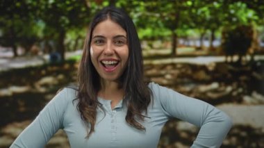 Yeşil ağaçlarla çevrili bir parkta gülümseyen genç İspanyol kadın neşeli bir ifade ve günlük kıyafetle güneşli havada eğleniyor..