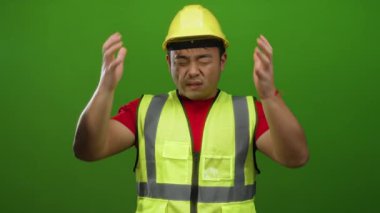Sarı kasklı ve yelekli bir adam, parlak yeşil arka planda yüzünü buruşturuyor, elleri başının üstünde stres yapıyor..