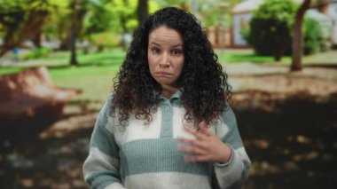 Genç, kıvırcık saçlı, yeşil ve banklarla çevrili çizgili bir süveter giymiş, düşünceli bir şekilde park ortamında jestler yapan İspanyol bir kadın, düşünceli bir ifade sergiliyor..