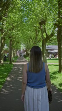 Güneşli yeşil parkta ağaçlarla kaplı yolda yürüyen bir kadın elinde örülmüş el çantasıyla mavi bluz ve beyaz etek giymiş kentsel bahçe ortamında canlı doğayla çevrili bir şekilde yürüyor..
