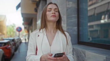 Beyaz ceketli kadın akıllı telefonu iki elinde tutuyor ve şehir caddesinde yan yan bakıyor; merak.