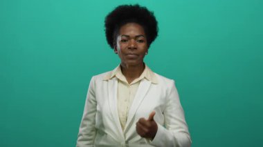 Beyaz takım elbiseli, afro saçlı bir kadın yeşil bir arkaplanı işaret ederek, dostça bir gülümsemeyle onaylama ve pozitifliği dile getiriyor..