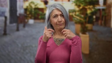 Gri saçlı yaşlı kadın canlı bir sokak ortamında duruyor, pembe bir kazak giyiyor, düşünceli bir ifade sergiliyor, kentsel yaşam tarzını yansıtıyor ve zarif bir şekilde yaşlanıyor..