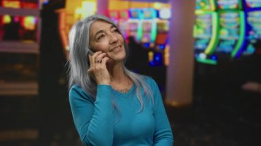 Gri saçlı bir kadın canlı bir kumarhanede kumar makineleriyle telefonda konuşuyor, canlı bir kapalı mekan atmosferi ve neşeli bir kıdemli etkileşimi sergiliyor..