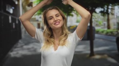 Bir şehir caddesinde elleri başının üstünde gülümseyen bir kadın, açık şehir ortamında gençlik ve neşe sergiliyor, sarı saçlarını ve beyaz özelliklerini ön plana çıkarıyor..