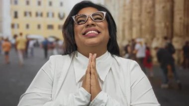 Kalabalık bir şehir caddesinde neşeli bir şekilde gülümseyen kadın, ellerini duayla kenetlenmiş, dış mekan ortamında gösteriş artı boyut ve kendine güvenen mutluluk..