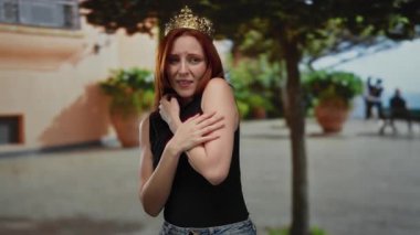 Kızıl saçlı ve tacı olan kadın ağaçlarla çevrili şehir caddesinde endişeli görünüyor..