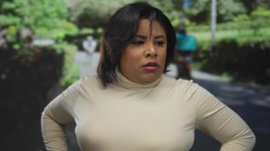 Bir park ortamında dışarıda duran bir kadın, arka planda yemyeşil yemyeşil, güneşli bir gökyüzünün altında, sakin, sakin, doğal bir çevre sergileyen düşünceli bir tavır sergiliyor..