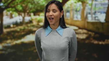 Arka planda ağaçlarla parkta dikilen bir kadın, dışarıda güneşli bir günde merak ve sürpriz sergiliyor..