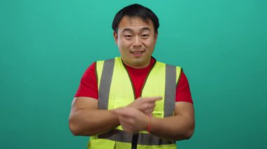 Güvenli yelek giyen genç adam, yalıtılmış yeşil arkaplanı işaret ederek yön ve güvence duygusunu gösteriyor..