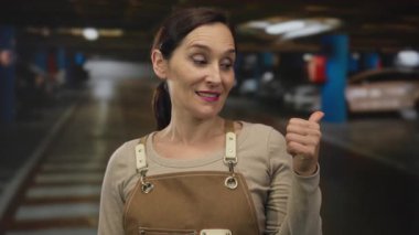 Önlüklü kadın bulanık arabaları ve arka planı olan loş ışıklı bir park yerinde olumlu duygu ve güveni ifade eden baş parmağını kaldırıyor..