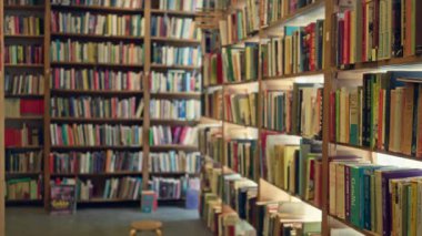 Renkli kitaplarla dolu raflarla bulanık kütüphane sahnesi. Kapalı bir okuma ortamı yaratıyor. Arka planda odakları bozulmuş ve ön planda küçük bir tabure var..