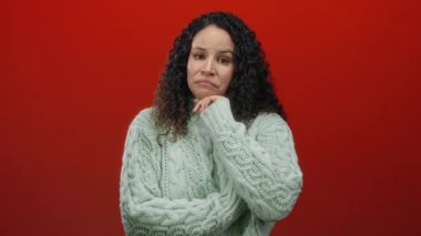 Yeşil kazaklı kadın canlı kırmızı bir arkaplanda düşünceli bir ifadeyle dikiliyor, kıvırcık saçlar sergiliyor ve izole bir arkaplanda sıradan bir moda sergiliyor..