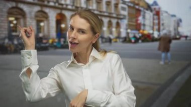 Kadın hareketli bir şehir caddesinde mutlu mesut işaret ediyor, şehir yaşam tarzını ve mimarisini sergiliyor, sarı saçı ve beyaz gömleğiyle açık hava sahnesine parlak bir kontrast ekliyor..