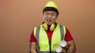 Kask ve güvenlik yeleği giymiş genç bir adam, elinde kahverengi bir arka plan, parlak kalpli profesyonelliği somutlaştıran planlar tutarken şakacı bir dille dilini uzatıyor..