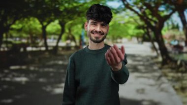 Güneşli bir günde, gölgeli ağaçların altında dostane ve davetkar bir atmosfer yaratan, yemyeşil bir park ortamında gülümseyen ve elini uzatan İspanyol bir adam..