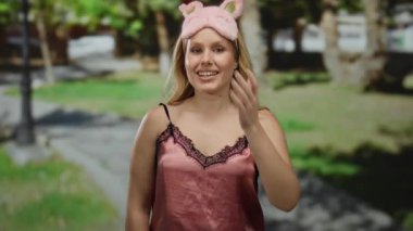 Pembe gecelikli, sarı saçlı, kedi kulaklı yeşil ağaçlarla çevrili güneşli bir parkta gülümseyen bir kadın..