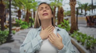 Güneşli bir parkta rahatsız hisseden bir kadın, etkileyici yüz hareketleriyle, gür palmiye ağaçları ile çevrili, açık hava ortamında..