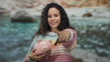 Sahilde elinde pembe bir kumbarayla duran, arka planda okyanus bulunan renkli süveter giyen bir kadın, dışarıda mali güvenini ve mutluluğunu gösteren kameraya bakıyor..