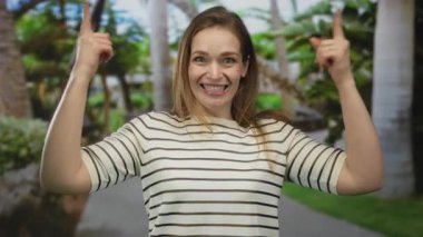 Açık ağızlı çizgili tişörtlü bir kadın gür ormanda parmaklarını yukarı doğru işaret ediyor..