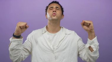 Genç adam kapalı gözlerle kutluyor ve mor bir arka plan önünde yumruklarını kaldırıyor, beyaz gömlek giyiyor ve güçlü duygular ifade ediyor..