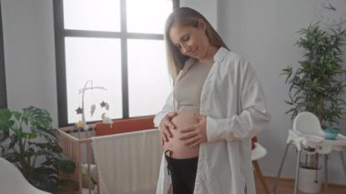 Bebek odasındaki rahat bir bebek odasında nazikçe gülümseyen hamile kadın, bebek mobilyasıyla çevrili, penceresinden güneş ışığı süzülen bir beşiğin yanında dururken anneliği kucaklıyor..