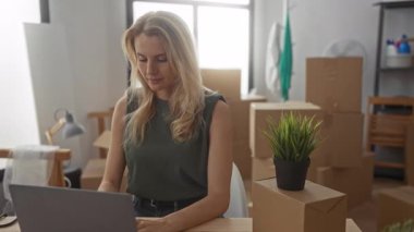 Oturma odasında dizüstü bilgisayarla çalışan bir kadın yeni evdeki kutuları taşıyarak, paketlenmemiş eşyalarla çevrili, yeni bir başlangıç ve taşınma senaryosunu temsil ediyor..