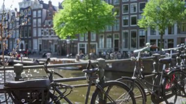 Amsterdam 'da bir kanal köprüsüne dayanan bisikletler arka planda bulanık, klasik Hollanda mimarisi ve güneşli gökyüzünün altında su yansımaları sergiliyor..