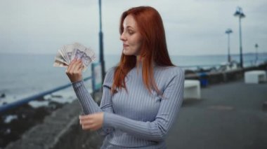 Okyanus ve gökyüzüyle çevrili, kendine güvenen bir gülümseme ile, deniz kenarında gezinirken elinde İsveç banknotları tutan kadın..