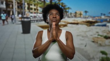 Deniz kenarındaki güneşli bir gezinti alanında huzur içinde meditasyon yapan bir kadın, dinginliği somutlaştırıyor ve canlı bir sahile odaklanıyor..