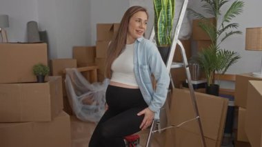 Yeni evindeki merdivenlerde oturan, modern bir oturma odasında kutuları ve bitkileri taşıyan, sarı saçlı hamile bir kadın heyecanlı bir yaşam sürecini ön plana çıkarıyor..