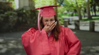 Kırmızı mezuniyet elbisesi giymiş genç bir kadın, güneşli bir parkta dikilirken, dışarıda kutlama anında rahatsızlığını dile getirirken endişeli görünüyor..