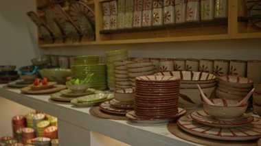 Bir çömlekçide düzgünce istiflenmiş renkli seramik bulaşık takımı, karmaşık desenli el yapımı çanaklar, tabaklar ve bardaklar sergileniyor..