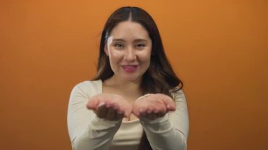 Uzun siyah saçlı genç İspanyol kadın stüdyoda açık avuçlar ve turuncu duvarlı nazik bir gülümseme sunar..