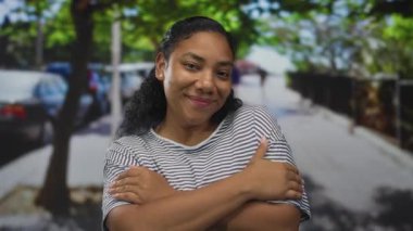 Çapraz kollu bir kadın güneşli bir şehir sokağında park etmiş arabaların ve ağaçların önünde kendine sarılıyor..