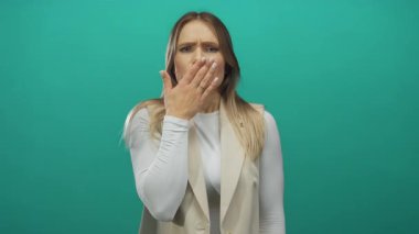 Sarışın genç bir kadın yeşil arka plana karşı şaşkınlıkla ağzını kapatıyor. Elleriyle dudaklarını kapayarak şaşkınlığını ifade ediyor. Duygularını ve merakını dile getiriyor..