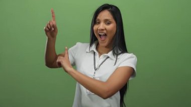 Gülümseyen ve yanlamasına gösteren beyaz gömlekli yeşil bir duvara yaslanmış bir kadın, dost canlısı ve davetkar bir atmosfer yayıyor..