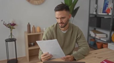 Evin oturma odasında sakallı genç bir adam, dizüstü bilgisayarından kağıda bakıyor, hayal kırıklığını ifade ediyor, basit dekor, bitkiler ve doğal ışık ile çevrili..